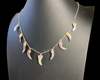 Puede incluir: Un collar de cadena plateada con siete colgantes en forma de cuerno. Cada colgante está hecho de piedra lisa de color violeta claro y blanco, con un alambre dorado enrollado en la parte superior donde se une a la cadena.