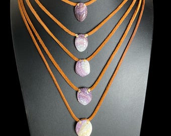 Puede incluir: Cinco collares superpuestos con cordones de ante marrón y colgantes ovalados de concha morada y blanca. Cada colgante muestra una superficie tallada y texturizada con un patrón distinto, suspendido del cordón por una pequeña anilla metálica.