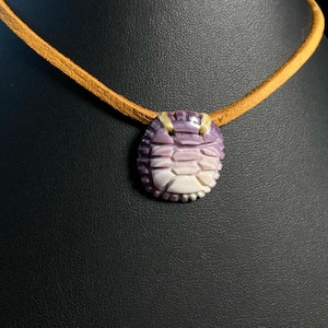 May include: A necklace featuring a carved, oval-shaped pendant made of purple and white shell material. The pendant has a segmented, ribbed texture and is suspended from a simple, light tan leather cord that loops through the top of the piece.