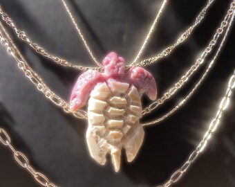 Puede incluir: Un collar de varias capas en tono plateado con un colgante de tortuga tallada. La tortuga tiene la cabeza y las aletas de color rosa con un caparazón de color crema. El collar consta de varias cadenas delicadas de diferentes longitudes y estilos.