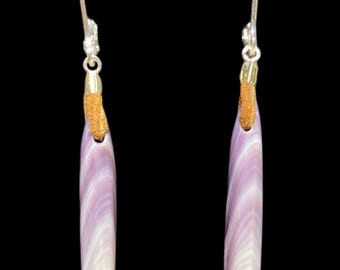 Puede incluir: Un par de pendientes colgantes con colgantes alargados en forma de lágrima con un patrón de remolinos en lavanda y blanco. Los colgantes están unidos a ganchos de metal plateado mediante un cordón texturizado de color canela en la parte superior.