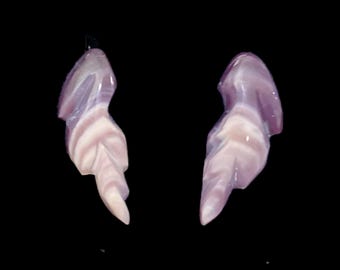 Puede incluir: Un par de pendientes formados por piedras talladas y pulidas con un acabado liso y brillante. Las piedras presentan un patrón de bandas superpuestas en tonos violeta claro, lavanda y blanco crema, estrechándose hacia una punta inferior.
