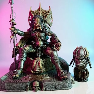 Könnte beinhalten: Eine detaillierte Predator-Statue, die auf einem Thron sitzt und einen Speer hält. Die Figur ist überwiegend silber, grün und braun, mit aufwendiger Rüstung und einem schädelverzierten Sockel. Eine kleinere Predator-Büste ist ebenfalls vorhanden.