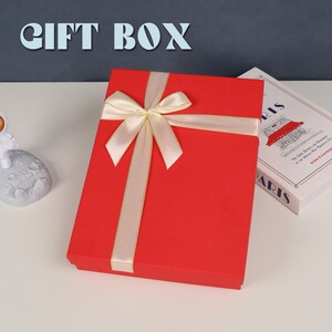 May include: A rectangular red gift box featuring a cream-colored satin ribbon tied in a bow across the top. The text GIFT BOX is displayed in large light blue letters above the box, which sits next to a book titled Paris.