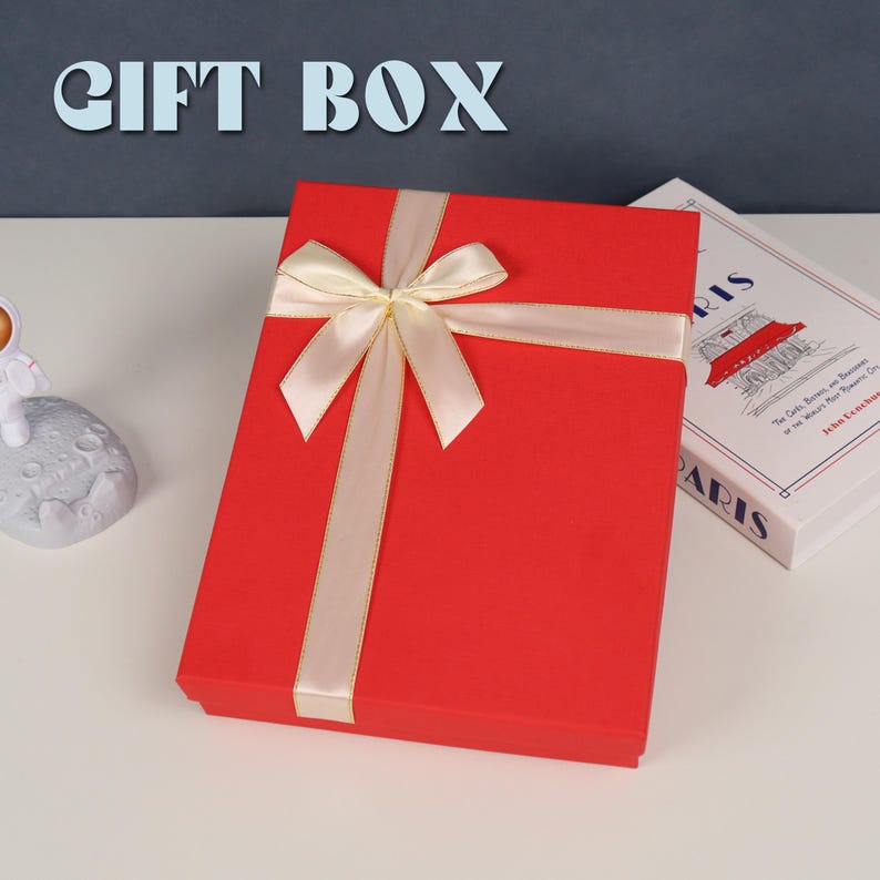 May include: A rectangular red gift box featuring a cream-colored satin ribbon tied in a bow across the top. The text GIFT BOX is displayed in large light blue letters above the box, which sits next to a book titled Paris.