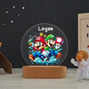 May include: A circular acrylic night light featuring a colorful graphic of Mario and Luigi splashing in water, with the name Logan printed in white at the top. The clear disc is mounted on a smooth, oval-shaped light-colored wooden base.