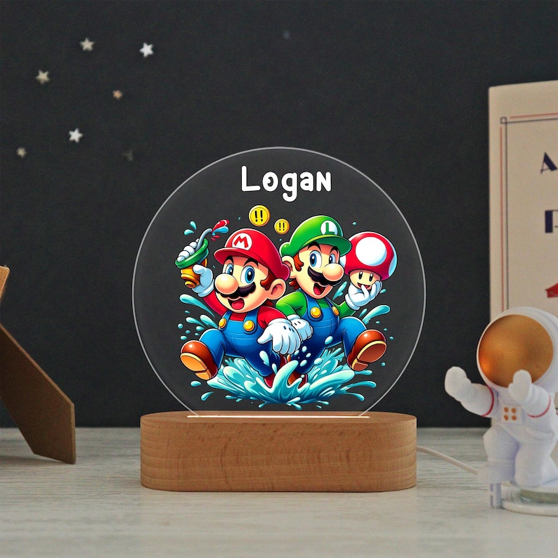 May include: A circular acrylic night light featuring a colorful graphic of Mario and Luigi splashing in water, with the name Logan printed in white at the top. The clear disc is mounted on a smooth, oval-shaped light-colored wooden base.