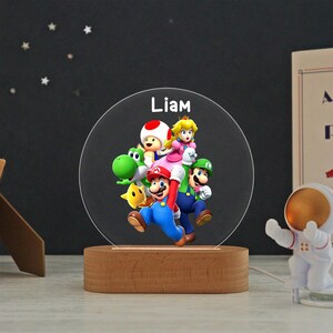 May include: A circular acrylic night light featuring a colorful graphic of Mario, Luigi, Princess Peach, Toad, Yoshi, and a yellow star. The name Liam is printed in white at the top. The light is mounted on a smooth, oval-shaped wooden base.