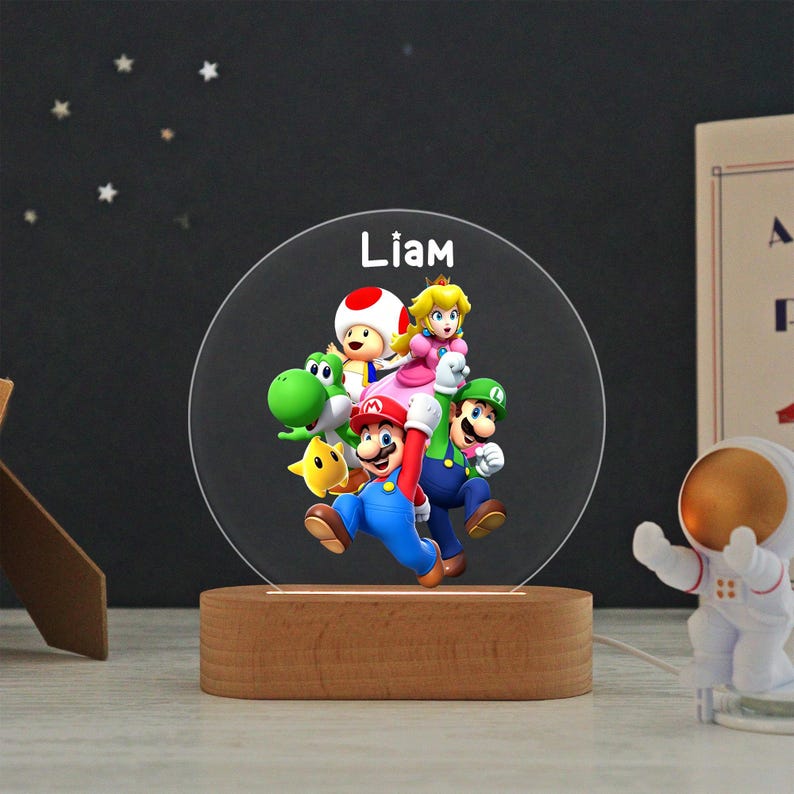 May include: A circular acrylic night light featuring a colorful graphic of Mario, Luigi, Princess Peach, Toad, Yoshi, and a yellow star. The name Liam is printed in white at the top. The light is mounted on a smooth, oval-shaped wooden base.