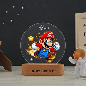 May include: A circular acrylic night light featuring a colorful graphic of Mario running, with the name Lucas printed above and the text Happy Birthday engraved on the oval wooden base. The design includes yellow stars and motion lines.