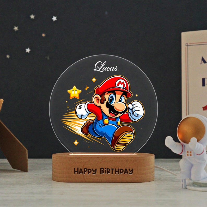 May include: A circular acrylic night light featuring a colorful graphic of Mario running, with the name Lucas printed above and the text Happy Birthday engraved on the oval wooden base. The design includes yellow stars and motion lines.