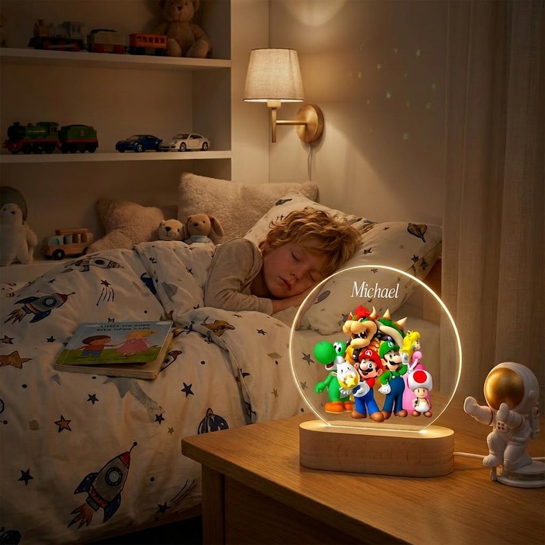May include: A circular acrylic night light with a wooden base featuring a colorful graphic of Mario, Luigi, Yoshi, Bowser, and Toad, with the name Michael printed at the top. The lamp emits a warm glow, illuminating the characters in a bedroom.