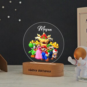 May include: A circular acrylic night light featuring a colorful graphic of Bowser, Yoshi, Princess Peach, Mario, Luigi, and Toad. The name Mason is printed above the characters. The light sits on a wooden base with the text HAPPY BIRTHDAY engraved.
