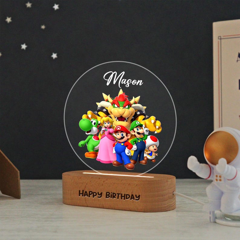May include: A circular acrylic night light featuring a colorful graphic of Bowser, Yoshi, Princess Peach, Mario, Luigi, and Toad. The name Mason is printed above the characters. The light sits on a wooden base with the text HAPPY BIRTHDAY engraved.