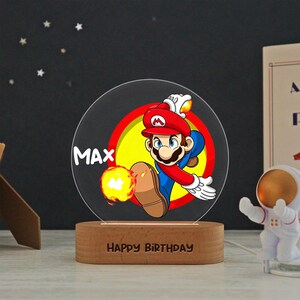 May include: A circular acrylic night light featuring a colorful graphic of Mario in a red hat and blue overalls, throwing a fireball. The name MAX is printed in white, and the wooden base is engraved with the text HAPPY BIRTHDAY in capital letters.