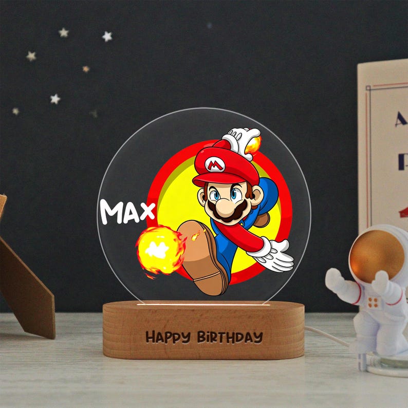 May include: A circular acrylic night light featuring a colorful graphic of Mario in a red hat and blue overalls, throwing a fireball. The name MAX is printed in white, and the wooden base is engraved with the text HAPPY BIRTHDAY in capital letters.