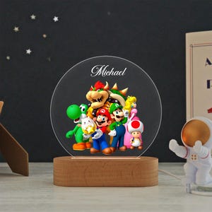 May include: A circular acrylic night light featuring a colorful graphic of Mario, Luigi, Princess Peach, Bowser, Yoshi, and Toad. The name Michael is printed in white script at the top. The light is mounted on a smooth, oval-shaped wooden base.