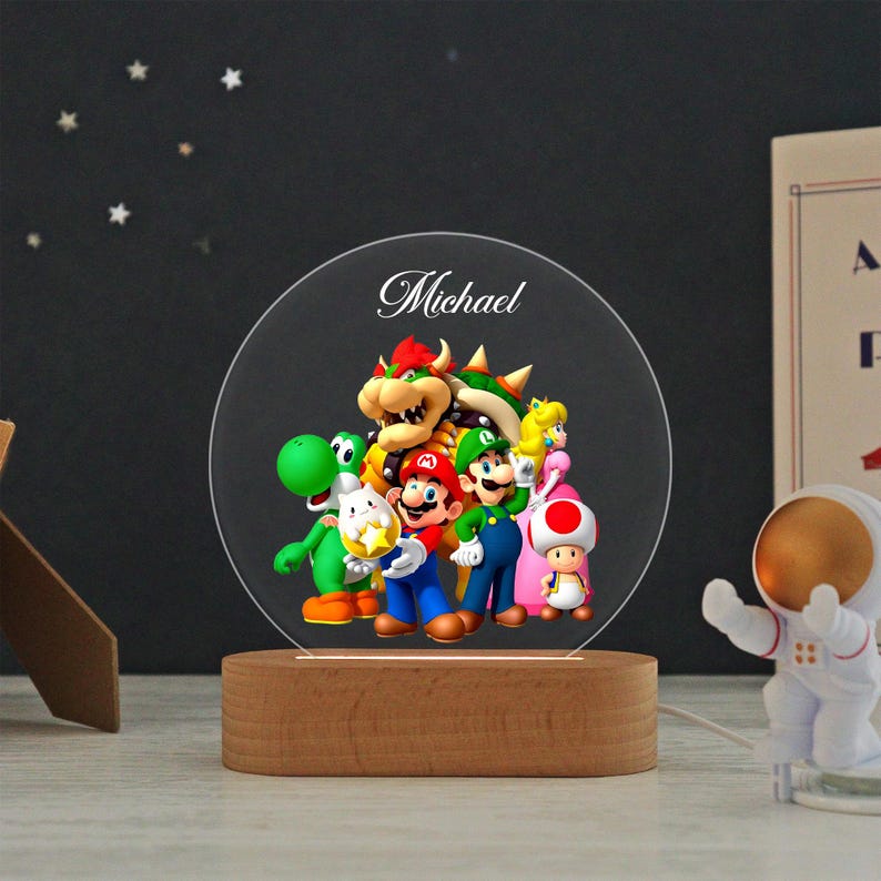 May include: A circular acrylic night light featuring a colorful graphic of Mario, Luigi, Princess Peach, Bowser, Yoshi, and Toad. The name Michael is printed in white script at the top. The light is mounted on a smooth, oval-shaped wooden base.