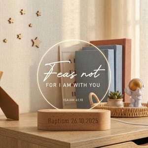 Puede incluir: Luz de noche LED circular con base de madera, que muestra el texto "Fear not, for I am with you" y la fecha "Bautismo 26.10.2025". La luz ilumina un círculo de acrílico transparente. Libros y adornos en el fondo.