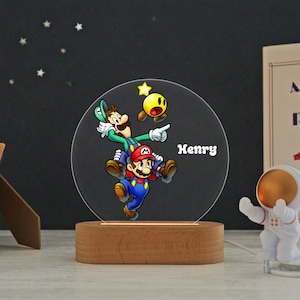 May include: A circular acrylic night light featuring a colorful graphic of Mario and Luigi jumping together with a yellow star. The name Henry is printed in white text on the right side. The light is mounted on a smooth, oval wooden base.