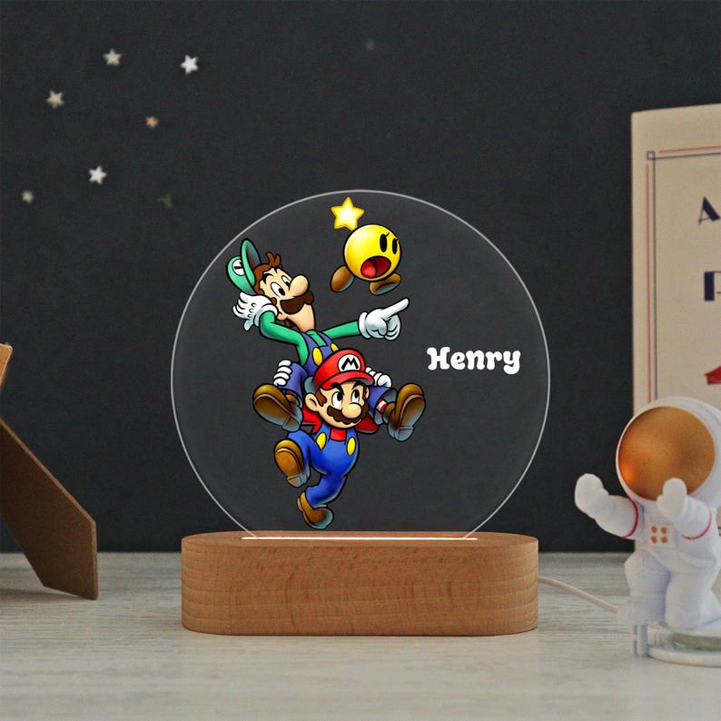 May include: A circular acrylic night light featuring a colorful graphic of Mario and Luigi jumping together with a yellow star. The name Henry is printed in white text on the right side. The light is mounted on a smooth, oval wooden base.