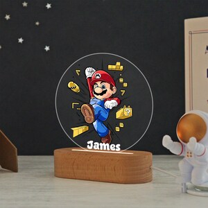 May include: A circular acrylic night light featuring a colorful illustration of Mario jumping, surrounded by yellow game blocks and a bullet bill. The name James is printed in white at the bottom. The light is set into a smooth, oval wooden base.
