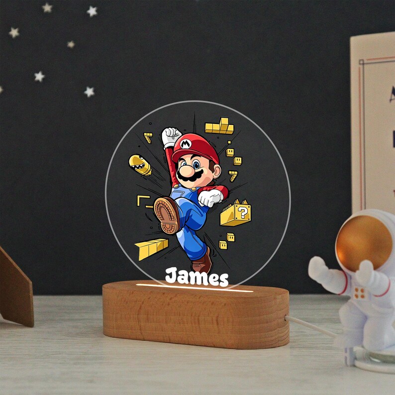 May include: A circular acrylic night light featuring a colorful illustration of Mario jumping, surrounded by yellow game blocks and a bullet bill. The name James is printed in white at the bottom. The light is set into a smooth, oval wooden base.