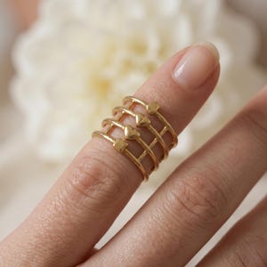 May include: A close-up of a gold-colored ring set. The ring set features multiple bands with heart and square accents. The rings are stacked on a finger, showcasing the design and detail of the jewelry.