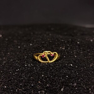 Puede incluir: Un anillo dorado con dos corazones entrelazados, cada uno adornado con una pequeña gema roja. El anillo se presenta sobre un fondo oscuro y texturizado, resaltando su intrincado diseño y cálido tono metálico. Una pieza de joyería romántica y elegante.