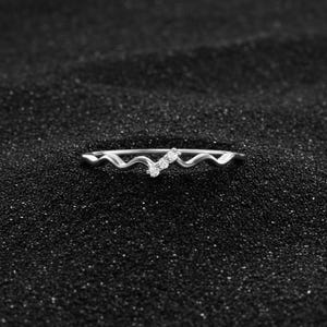 Puede incluir: Un anillo plateado con un diseño ondulado, adornado con tres pequeñas piedras preciosas transparentes. El anillo se encuentra sobre un fondo oscuro y texturizado, creando un contraste que resalta los detalles de la joya. El diseño es simple y elegante.