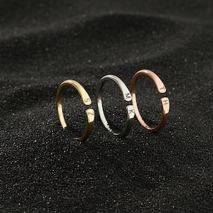 Puede incluir: Tres anillos abiertos en oro, plata y oro rosa. Cada anillo presenta una inicial: "SZ", "MK" y "TH". Los anillos se exhiben sobre una superficie oscura y texturizada, mostrando su diseño minimalista y toque personalizado.