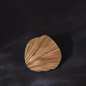 May include: A golden-coloured, fan-shaped decorative object with a textured surface. The object has a series of parallel lines radiating from a central point. The background is a dark, textured surface.
