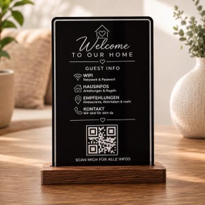 May include: A rectangular black sign with white text and icons, featuring the words Welcome To Our Home, Guest Info, and a QR code. The sign is mounted on a rectangular wooden base with a visible wood grain texture, displaying information in English and German.