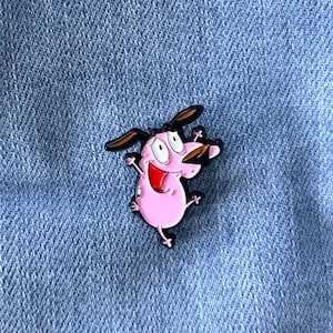Könnte beinhalten: Ein rosa Emaille-Pin von Courage, dem feigen Hund, mit schwarzen Akzenten, befestigt an einem hellblauen Jeansstoff. Der Pin zeigt den Cartoon-Hund mit überraschtem Gesichtsausdruck.