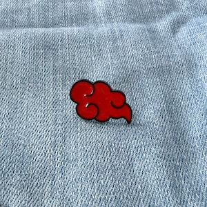 Puede incluir: Un pin de esmalte rojo en forma de nube con contorno negro, colocado sobre la mezclilla azul claro de un bolsillo de jean. El pin es un accesorio vibrante, que añade un toque de color a la prenda.