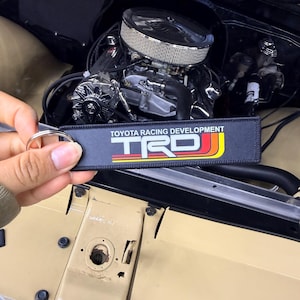 May include: Black keychain with the text "TOYOTA RACING DEVELOPMENT TRD" in white, yellow, and red. The keychain is held in front of a car engine with chrome and silver components. The car's hood is beige.