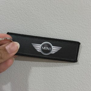 May include: Black rectangular keychain tag featuring the Mini logo. The logo is a silver emblem with the word "MINI" in the centre. The tag has a silver key ring attached.