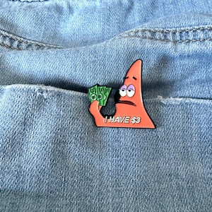 May include: A pink Patrick Star-shaped pin holding green money, with the text "I HAVE $3". The pin is set against a blue denim background, likely a pocket. The pin has black outlines and a bored expression.
