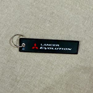 May include: Black rectangular keychain with the words "Lancer Evolution" in white text. The keychain also features a red Mitsubishi logo. A silver key ring is attached to the top left corner. The background is a tan fabric.