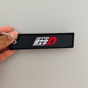 Puede incluir: Llavero rectangular negro con el texto "INITIAL 頭文字D" en blanco y rojo. El llavero tiene un anillo de metal plateado. El fondo es gris claro.