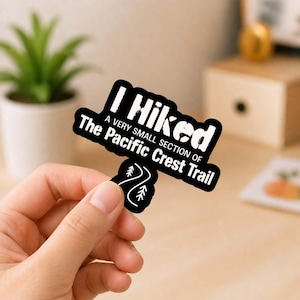 May include: A black die-cut sticker with white text that reads I Hiked A VERY SMALL SECTION OF The Pacific Crest Trail, featuring a small white graphic of a winding path with two pine trees at the bottom of the sticker design.