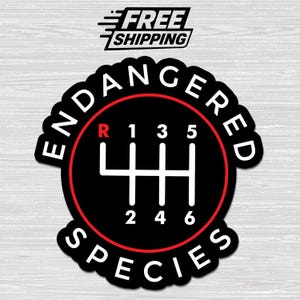 May include: A circular sticker featuring a black background with a white manual transmission gear shift pattern. The letter R is in red, and the numbers 1 through 6 are in white. The text ENDANGERED SPECIES is printed in white along the border.