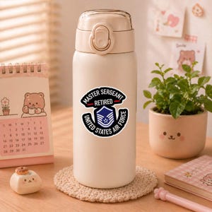 May include: A beige insulated water bottle with a silver lid and a sticker that reads "MASTER SERGEANT RETIRED UNITED STATES AIR FORCE". The bottle is sitting on a beige coaster. A calendar and small potted plant are in the background.