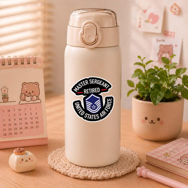 May include: A beige insulated water bottle with a silver lid and a sticker that reads "MASTER SERGEANT RETIRED UNITED STATES AIR FORCE". The bottle is sitting on a beige coaster. A calendar and small potted plant are in the background.