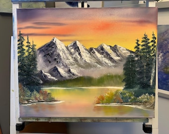 May include: An oil painting on canvas depicting a mountain range with snow-capped peaks under a sunset sky. A calm lake reflects the orange and yellow sky, surrounded by dark green pine trees and colorful autumn bushes along the shoreline.