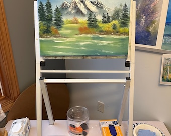 May include: A white metal easel holds a landscape oil painting of snow-capped mountains, evergreen trees, and a calm lake. Below the painting, a shelf holds a clear plastic container of sponges, a box of blue nitrile gloves, and white plastic clips.