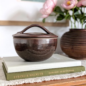 May include: A dark brown, glazed ceramic bowl with a matching lid featuring a central arched handle. The bowl has a smooth, rounded body and sits atop two stacked hardcover books, one light gray and one olive green with gold lettering.