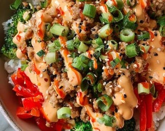 May include: A close-up shot of a bowl filled with a colorful and appetizing meal. The dish features white rice, topped with cooked ground meat, vibrant red bell peppers, green broccoli florets, and sliced green onions. A creamy orange sauce and sesame seeds complete the dish.