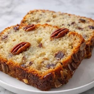 Könnte beinhalten: Zwei Scheiben Bananen-Nuss-Brot auf einem weißen Teller. Das Brot hat eine goldbraune Kruste und ist mit Pekannüssen und Nüssen bestückt. Das Innere des Brotes ist feucht und hell.