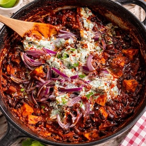 Op de afbeelding: Een close-up van een gietijzeren pot gevuld met chili. De chili bevat zwarte bonen, stukjes zoete aardappel en rode ui, gegarneerd met een romige, lichtgroene saus. Een houten lepel rust in de chili.